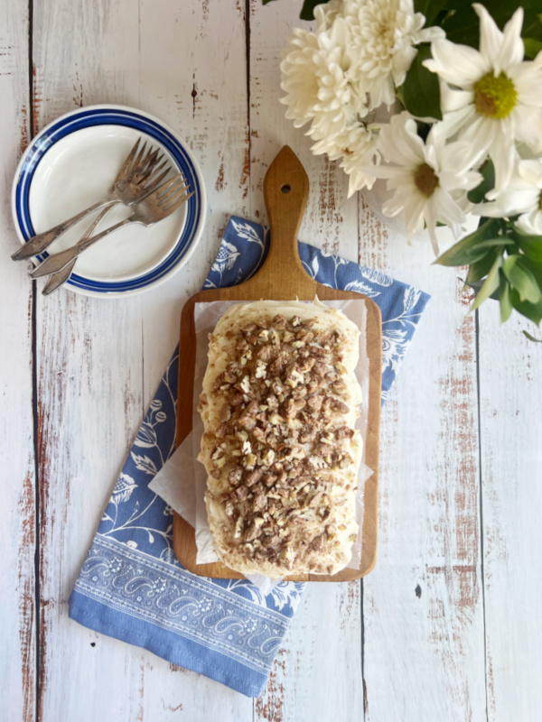 Cannot banana bread with cream cheese frosting on a cutting board with a vase of white flowers, a plate white with blue rim sitting on a blue and white tea towel. 