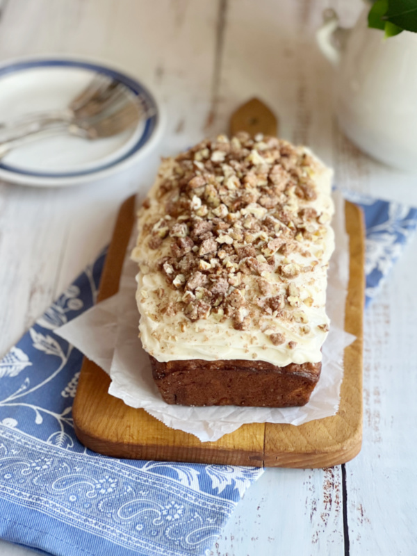 Cream cheese frosted banana bread with candied pecans sitting on a cutting board atop a blue and white tea towel.