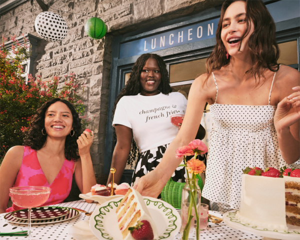 Three women around a white dress with strawberries with one handing a piece to the others, Kate Spade NY x Target.