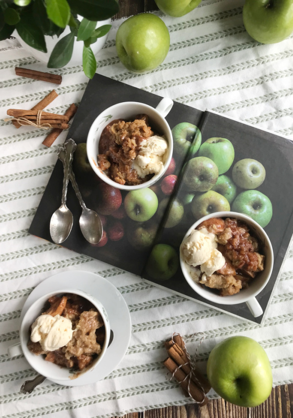 Ina Garten's apple crisp in 3 mugs sitting on an open book ov apples. apple Crisp