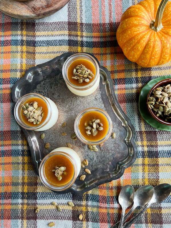 Panna Cotta with pumpkin seed brittle on a silver tray with an orange pumpkin and a bowl of pumpkin seeds sitting on an orange plaid blanket. 