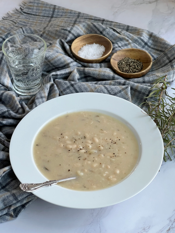 Barefoot Contessa Rosemary white bean soup in a white bowl. with a spoon sitting on a gray plaid throw. with a glass of sparkling water.