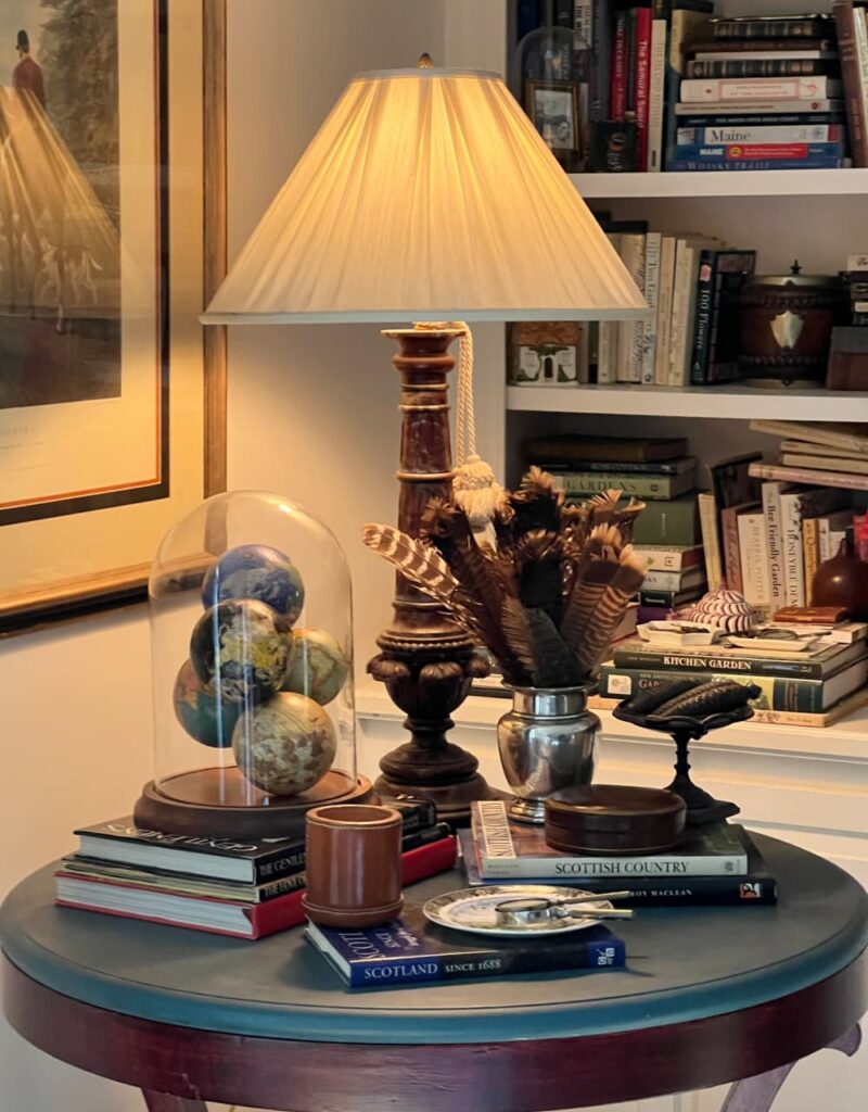 Maximalist Home Decor Table piled with books, a lamp, leather boxes, a silver pot of feathers.