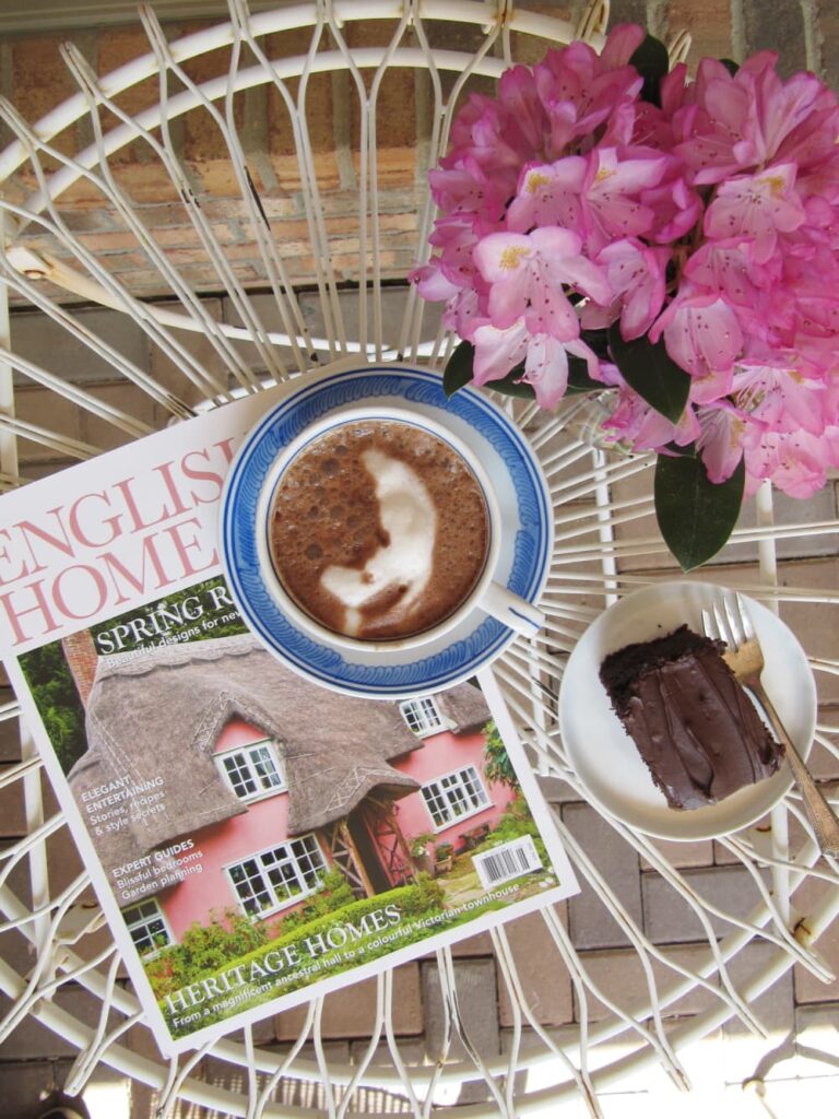 White iron table with a magazine, a piece of cake, cup of hot chocolate and a vase of fuchsia flowers.