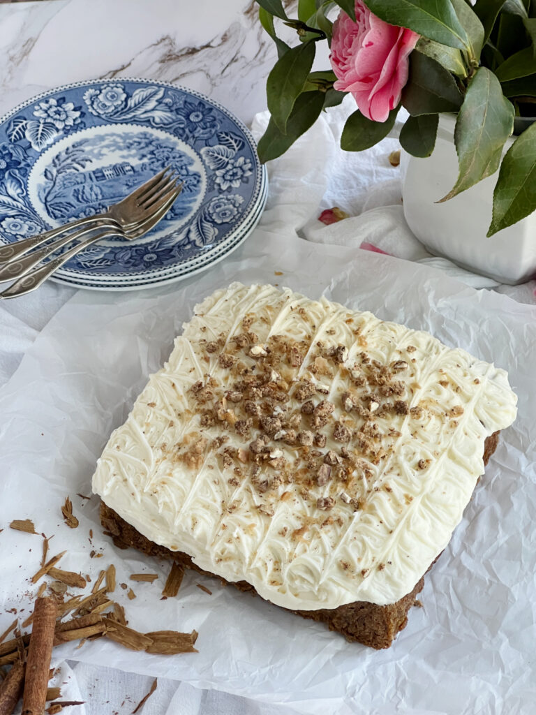 carrot cake with cream cheese frosting a stack of blue and white plates with forks and a white vase with pink camellia flowers.