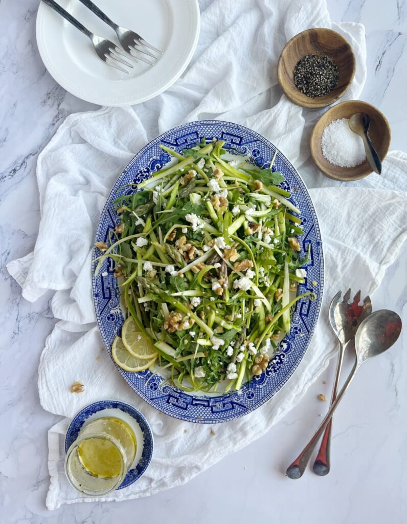 haved asparagus salad with feta cheese and lemon slices on a blue and white platter with salt and pepper in wooden bowls.