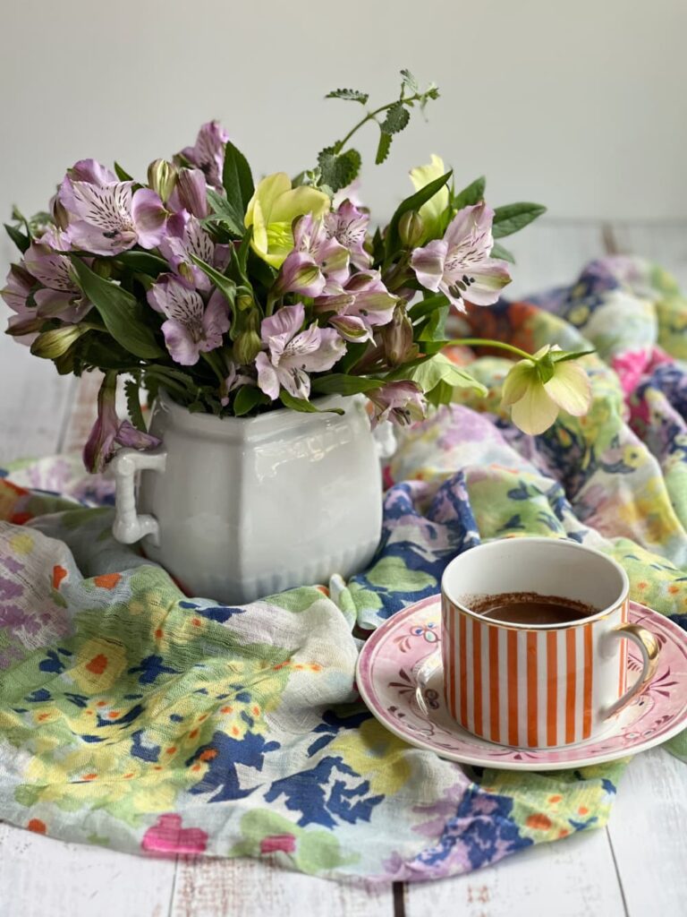 Spring flowers in a white ironstone vase with an orange and white cup of drinking chocolate.