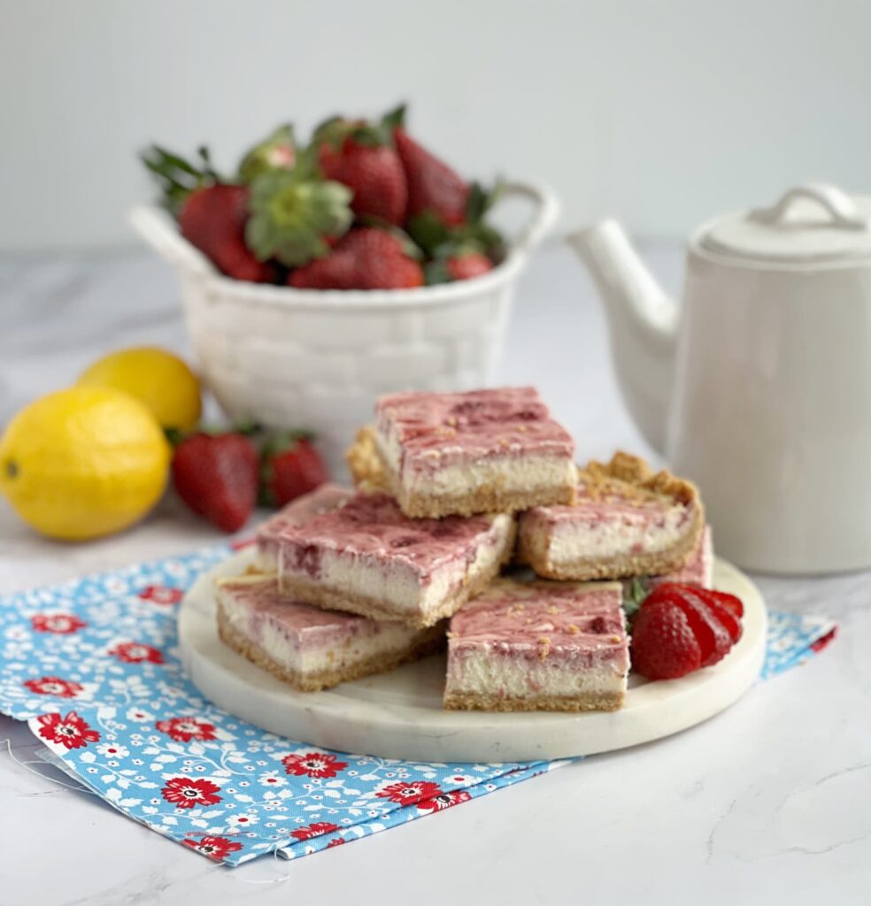 Strawberry Cheesecake Bars stacked on a white platter with a bowl of fresh strawberries and lemons