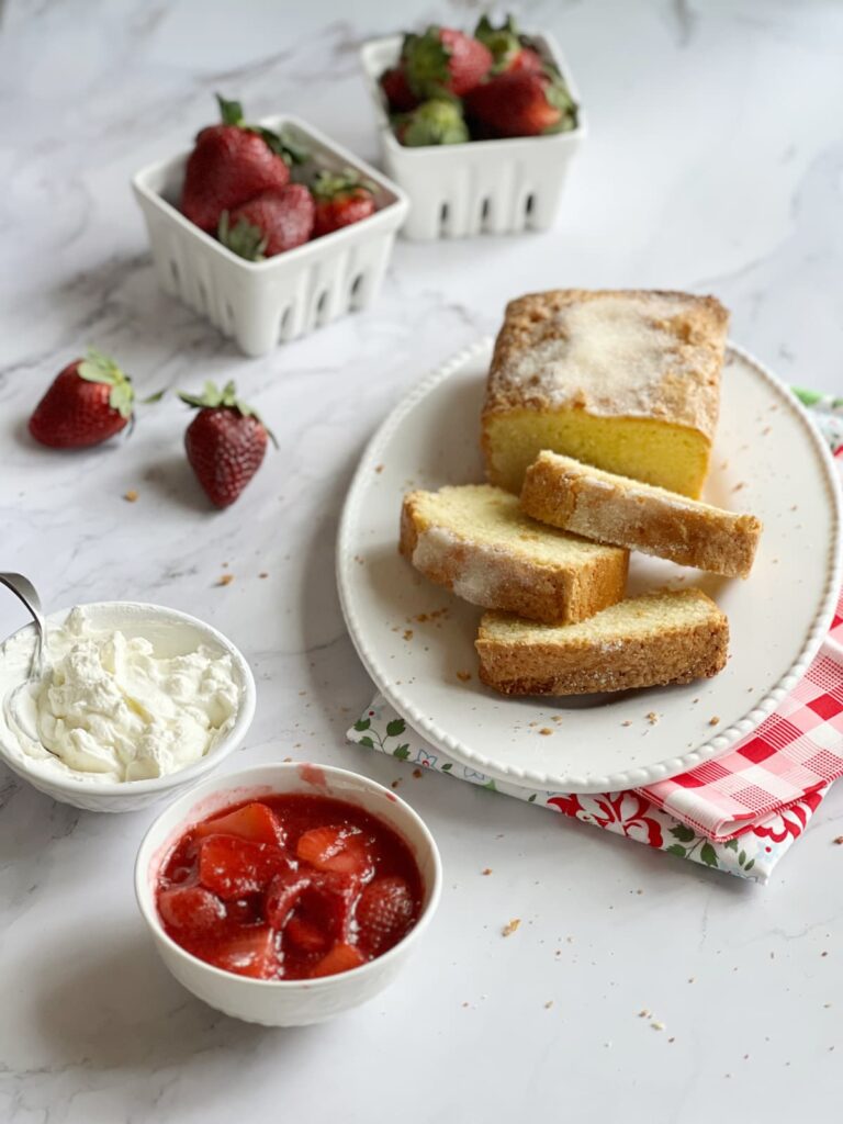pound cake on a white platter with a bowl of strawberry compote and a bowl of whipped cream.