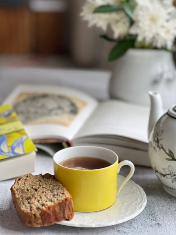 Recipes for entertaining — tea-in-a-yellow-cup-with-a-slice-of-banana-bread-and-2-books-with-a-teapot-and-a-vase-of-flowers.-