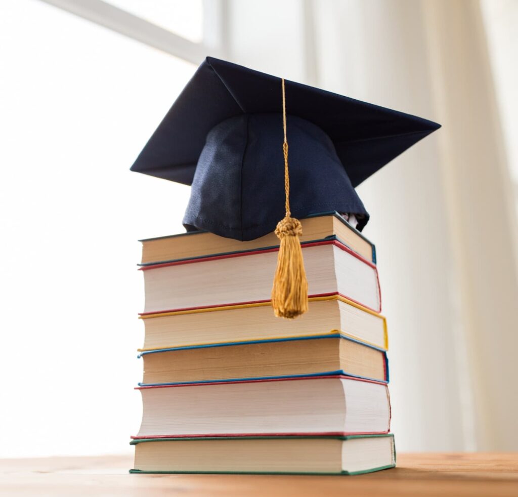 A stack of books with a graduation cap on a sun-lit table