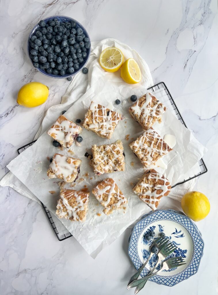 Sliced blueberry coffee cake with crumble topping and a drizzle of icing alongside a bowl of blueberries and some lemons.