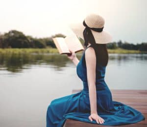 woman-wearing-a-blue-dress-and-a-straw-hat-sitting-on-the-edge-of-a-wood-pier-reading-a-book-by-water