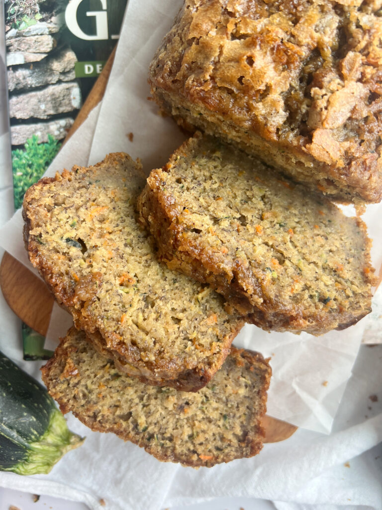 carrot zucchini banana bread sliced on a wood cutting board with a zucchini laying on the side